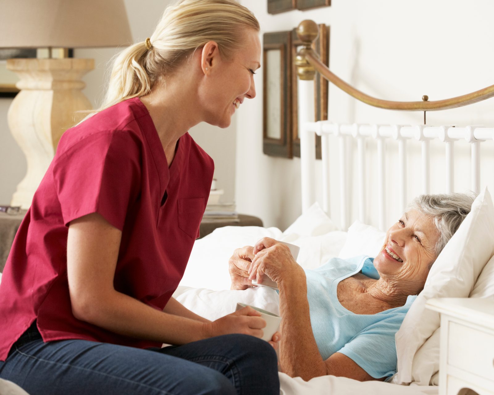 A caregiver sitting at the bedside sharing a warm drink and a laugh with an older woman at home
