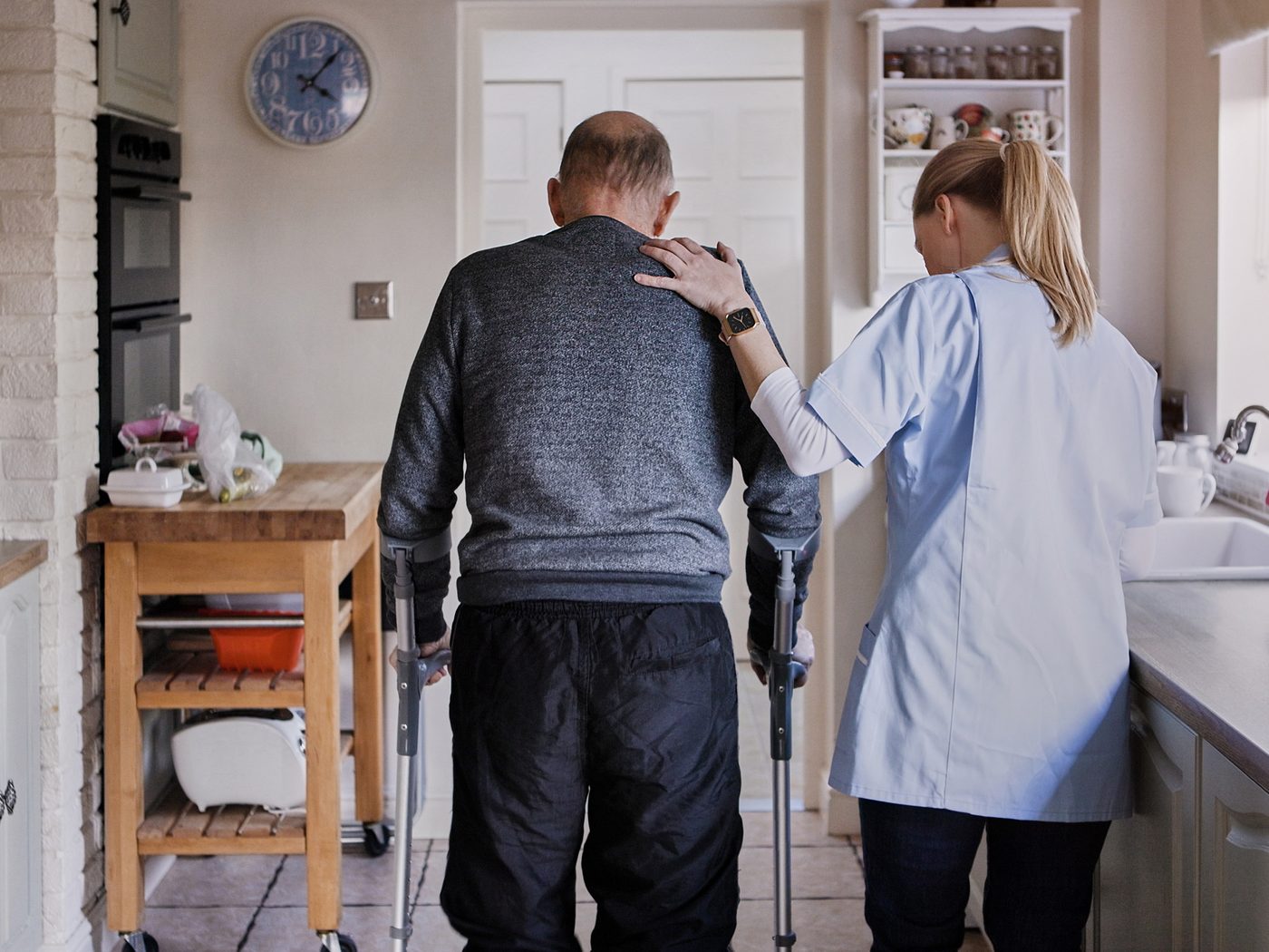 A SteadiCare caregiver steadying a man walking with crutches through his kitchen